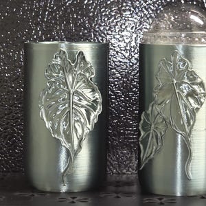 May include: Two cylindrical silver-toned containers with a brushed metallic finish. The left container features a single embossed leaf design, while the right container displays two overlapping embossed leaves and a clear, rounded dome-shaped lid.