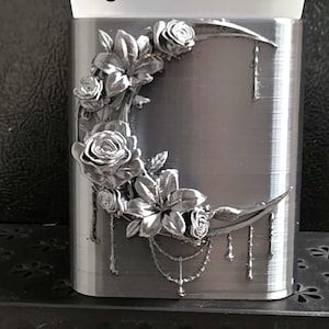 May include: A rectangular silver-colored container featuring a raised crescent moon design adorned with sculpted roses and lilies. The letter C is printed in black near the top. Delicate dangling chain-like elements hang from the moon motif.