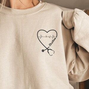 May include: A beige sweatshirt with a black heart graphic and a stethoscope. The heart graphic has a black EKG line and the text "Christina" inside the heart.