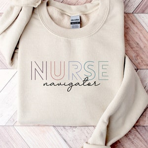 May include: A beige sweatshirt with the text "NURSE navigator" in colorful outlined letters. The text is in a rainbow color scheme.