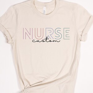 May include: A beige t-shirt with the word "NURSE" in rainbow colors and the word "custom" written underneath.