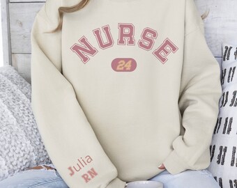 May include: A light beige sweatshirt with the word "NURSE" in pink lettering with a black outline. The number "24" is in a yellow oval. The left sleeve has the words "Julia RN" in pink lettering with a black outline.