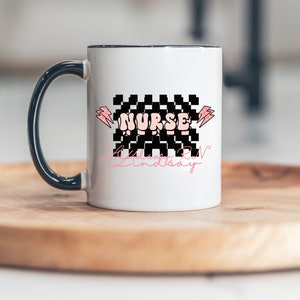 May include: White ceramic mug with a black handle. The mug has a black and white checkered design with the word "NURSE" in pink letters. The text "Lindsay RN" is written in pink cursive below the checkered design.