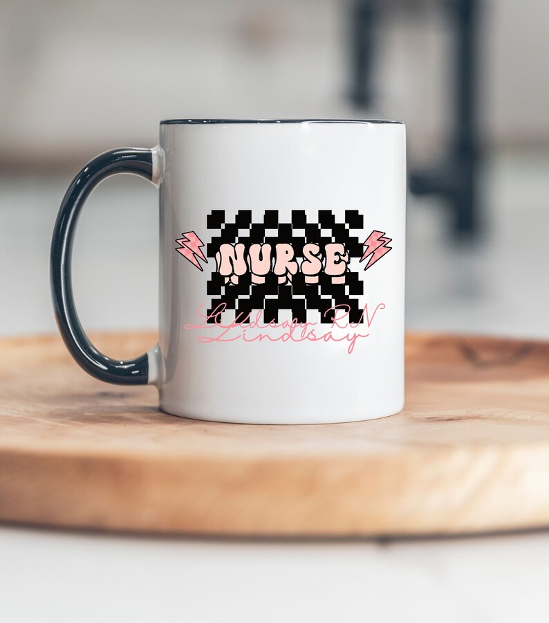 May include: White ceramic mug with a black handle. The mug has a black and white checkered design with the word "NURSE" in pink letters. The text "Lindsay RN" is written in pink cursive below the checkered design.