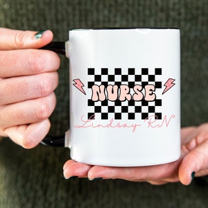 May include: White ceramic mug with a black rim and handle. The mug features a black and white checkered pattern with a pink "NURSE" graphic and lightning bolts. The text "Lindsay RN" is written in cursive below the checkered pattern.