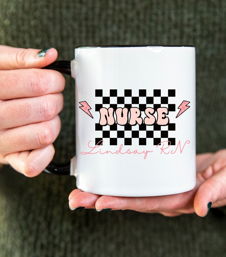May include: White ceramic mug with a black rim and handle. The mug features a black and white checkered pattern with a pink "NURSE" graphic and lightning bolts. The text "Lindsay RN" is written in cursive below the checkered pattern.