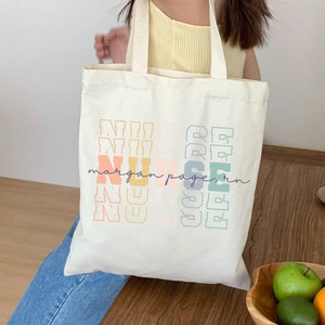 May include: A white canvas tote bag with the word "NURSE" printed in rainbow colors. The text "morgan page, rn" is printed below the word "NURSE" in a smaller font.