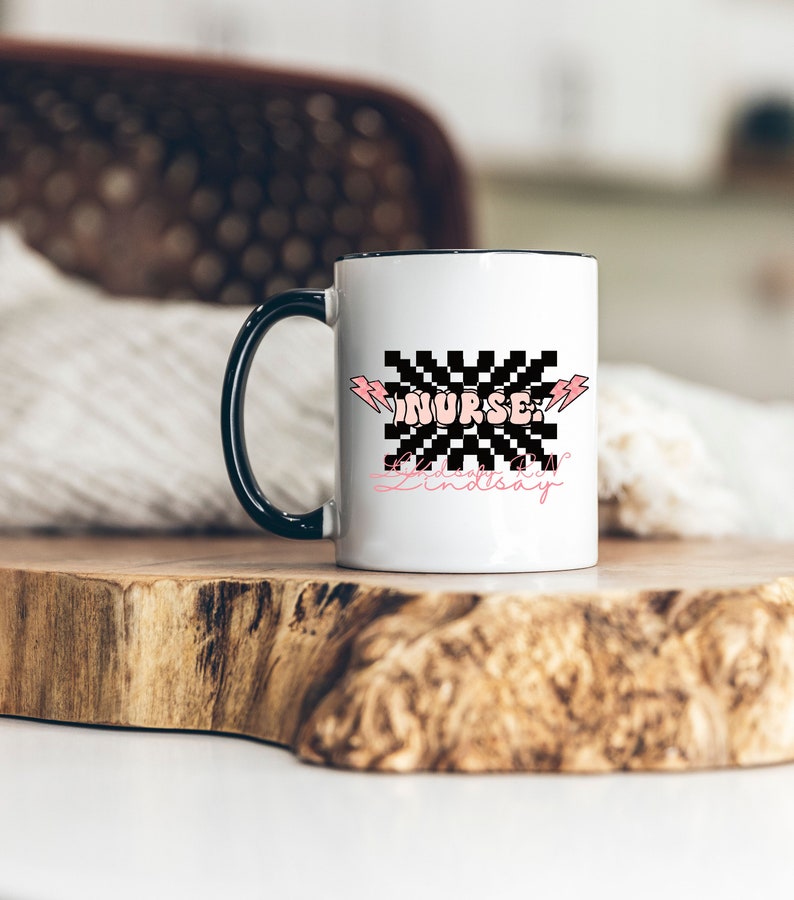 May include: White ceramic mug with a black handle. The mug has a black and white checkered design with the word "NURSE" in pink letters. The text "Always be kind" and "Lindsay" are written in pink cursive below the checkered design.