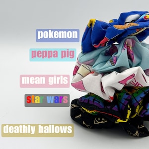 May include: A stack of five colorful scrunchies with different cartoon characters and logos. The scrunchies are in the following order from top to bottom: Pokemon, Peppa Pig, Mean Girls, Star Wars, and Deathly Hallows.