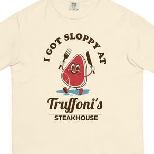 May include: A cream-colored short-sleeve t-shirt featuring a graphic of a cartoon steak character holding a fork and knife. The text reads I GOT SLOPPY AT Truffoni's STEAKHOUSE in brown lettering. The steak is red with white details.