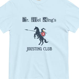 May include: Pale blue t-shirt with the words "Dr. Mel King's Jousting Club". The design shows a knight on a horse with a lance and shield. The shield has a red cross.