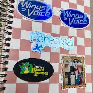 May include: A notebook with a spiral binding and a pink and white chequered pattern. Several stickers are attached, including two with "Wings of Voice" text, one with "The Rehearsal" text and an aeroplane graphic, and one with "Nate's Lizard Lounge" text.