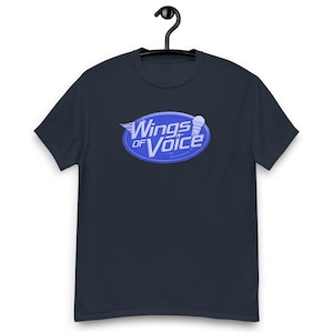 Pode incluir: Camiseta azul marinho com o texto "Wings of Voice" em um design oval azul e branco. O design inclui asas e um microfone. A camiseta está pendurada em um cabide preto.