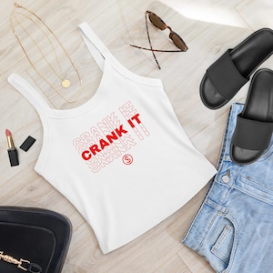May include: White tank top with the text "CRANK IT" in red. The top is paired with denim shorts, black sandals, and gold jewelry. Sunglasses and lipstick are also visible.