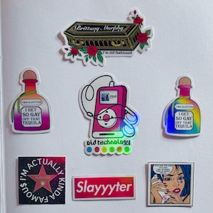 May include: A collection of colorful stickers with various designs. One sticker features a coffin with "Brittany Murphy" and "I'm old fashioned." Other stickers include a pink MP3 player, tequila bottles with text, a star, and the word "Slayyyter."
