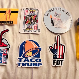 以下が含まれることがあります： ステッカーとライターのコレクション。「KILL BILLIONAIRES」、トランプ、蝶ネクタイをした猫、氷のカップ、「TACO TRUMP」のタコベルのベル、アメリカ国旗と「FDT」の手など、ステッカーが含まれています。