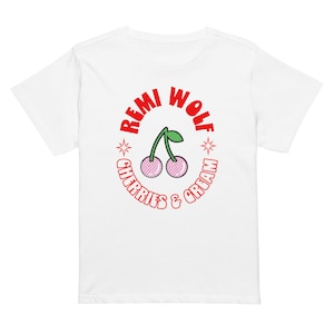 Puede incluir: Camiseta blanca con un diseño circular rojo. El diseño presenta el texto "REMI WOLF" y "CHERRIES & CREAM" alrededor de un par de cerezas rosas con tallos y hojas verdes. El diseño también incluye estrellas.