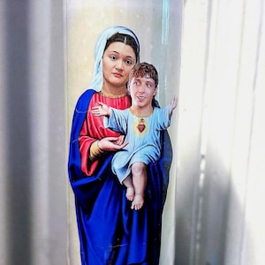 Könnte beinhalten: Eine Glaskerze mit einem religiösen Bild einer Frau und eines Kindes. Die Frau trägt einen blauen Schleier und rote und blaue Gewänder. Das Kind hat ein rotes Herz auf der Brust. Die Kerze ist mit "Saint Fuckleberry" beschriftet.
