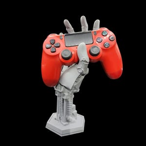 May include: A 3D printed robotic hand holding a red video game controller. The hand is attached to a gray base with a hexagonal shape.