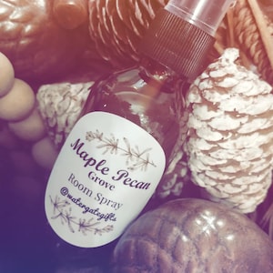 Puede incluir: Una botella de vidrio marrón con una etiqueta blanca que dice "Maple Pecan Grove Room Spray" y "@watergategifts". La botella está sobre una superficie marrón con piñas.