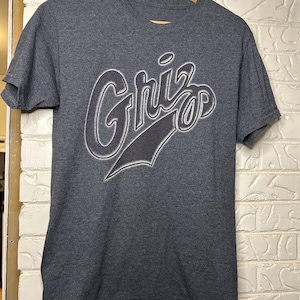 Puede incluir: Una camiseta gris oscuro con la palabra "Griz" en una fuente estilizada. La palabra está delineada en blanco con una sombra gris oscuro. La camiseta está colgada en una percha de madera contra una pared de ladrillos blancos.