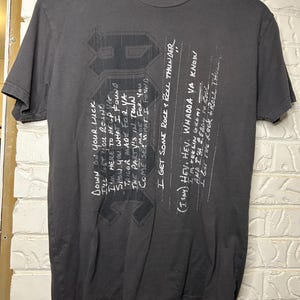 Op de afbeelding: Donkergrijs t-shirt met een grafisch ontwerp en handgeschreven tekst. De tekst bevat zinnen als "Down on your luck" en "I get some rock & roll thunder." Het t-shirt hangt aan een houten hanger.