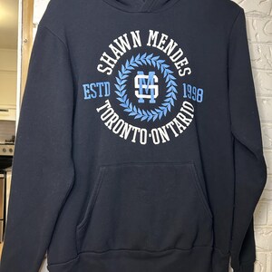 Könnte beinhalten: Ein marineblauer Kapuzenpullover mit einer Vordertasche. Der Pullover hat eine kreisförmige Grafik mit dem Text "SHAWN MENDES ESTD 1998 TORONTO ONTARIO" in Weiß und Hellblau. Die Grafik enthält ein stilisiertes "SM"-Monogramm.