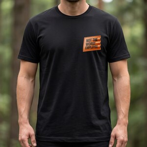 May include: A black short-sleeved t-shirt featuring a rectangular orange patch on the left chest. The patch contains the black text "NOT THE WORST EMPLOYEE" in a bold, sans-serif font. The shirt has a crew neckline and a standard fit.