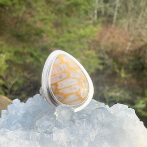 May include: A silver ring with a teardrop-shaped stone. The stone is a light yellow and white with a patterned design. The ring is sitting on a bed of blue crystals.