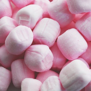 May include: A collection of small, square-shaped, light pink sweets with rounded edges. Each piece has a smooth, matte surface texture and a uniform pastel pink colour, densely packed together to fill the entire frame of the image.