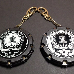 May include: Two black and white disc golf keychains with gold-colored clasps. Each keychain features a skull design with a disc golf basket graphic in the center. Numbers 0-18 are around the edge of the keychain.