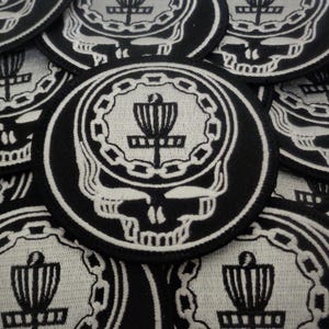 May include: A close-up of several black and white embroidered patches featuring a skull with a disc golf disc inside. The patches are circular and have a white outline.