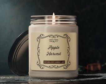 Puede incluir: Una vela blanca encendida en un tarro de cristal con tapa negra. La etiqueta dice "Apple Harvest" en escritura elegante, con "Natural Soy Candle - 9oz" debajo. El fondo es oscuro.