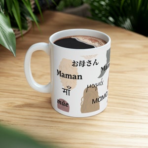 May include: White ceramic mug filled with dark coffee. The mug features the word "Maman" in multiple languages, including Japanese, Hindi, and Greek, along with illustrations of hair. The mug sits on a wooden surface.
