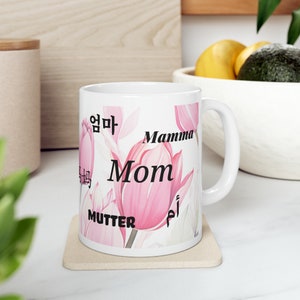 May include: White ceramic mug with a floral design featuring pink tulips. The mug has the word "Mom" in black lettering and the word "Mamma" in black lettering. The words "Mutter", "엄마", and "أم" are also written on the mug in black lettering.