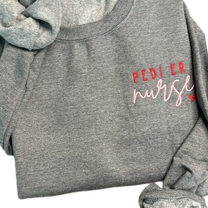 May include: A heather gray sweatshirt with the text "PEDI ER nurse" embroidered in red and pink on the left side of the chest.
