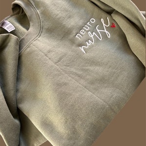 Puede incluir: Una sudadera verde con el texto "neuro nurse" bordado en blanco con un peque&ntilde;o coraz&oacute;n rojo.
