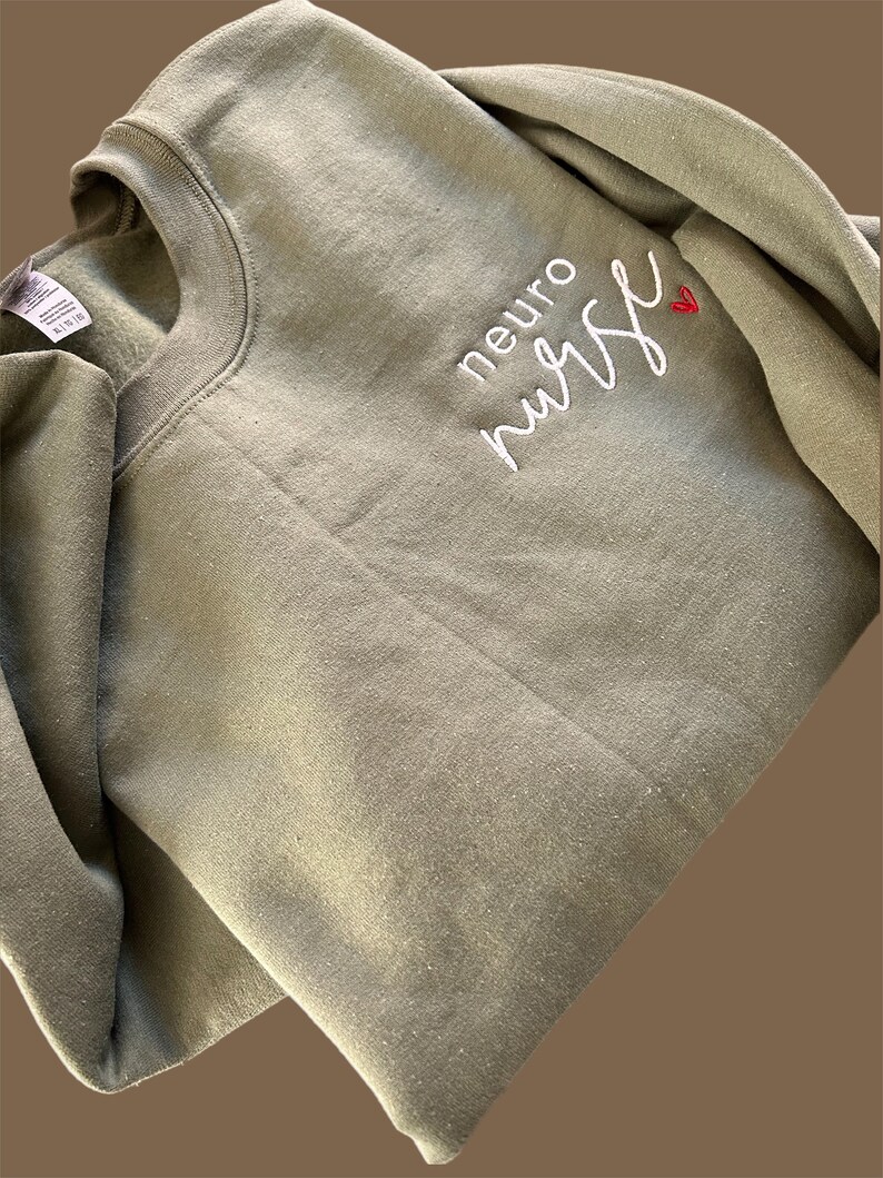 Puede incluir: Una sudadera verde con el texto "neuro nurse" bordado en blanco con un peque&ntilde;o coraz&oacute;n rojo.