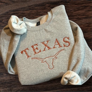 May include: A heather gray sweatshirt with the word "TEXAS" embroidered in burnt orange, along with a longhorn symbol. The sleeves are cuffed, revealing a white interior. The sweatshirt is folded on a wooden surface.