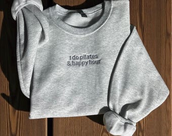 Può includere: Una felpa grigio mélange con la scritta ricamata "I do pilates & happy hour". La parte superiore a girocollo a maniche lunghe è piegata su una superficie di legno. La felpa è in stile casual.