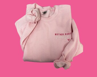 Può includere: Una felpa rosa con il testo "MOTHER BABY NURSE" ricamato in bianco con un cuore rosa.