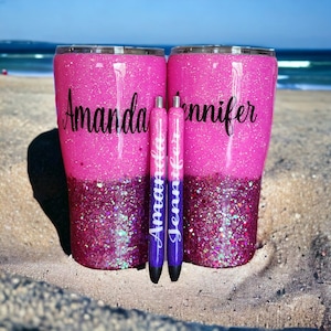 May include: Two pink and purple glitter tumblers with the names "Amanda" and "Jennifer" written on them. The tumblers are sitting on a sandy beach with two purple pens in front of them.