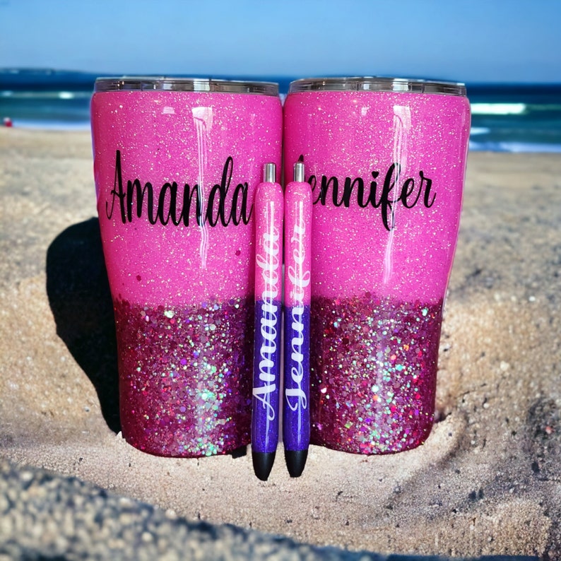 May include: Two pink and purple glitter tumblers with the names "Amanda" and "Jennifer" written on them. The tumblers are sitting on a sandy beach with two purple pens in front of them.