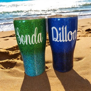 May include: Two personalized glitter tumblers with ombre colors. The tumbler on the left is green and blue with the name "Sonda" on it. The tumbler on the right is blue with the name "Dillon" on it.