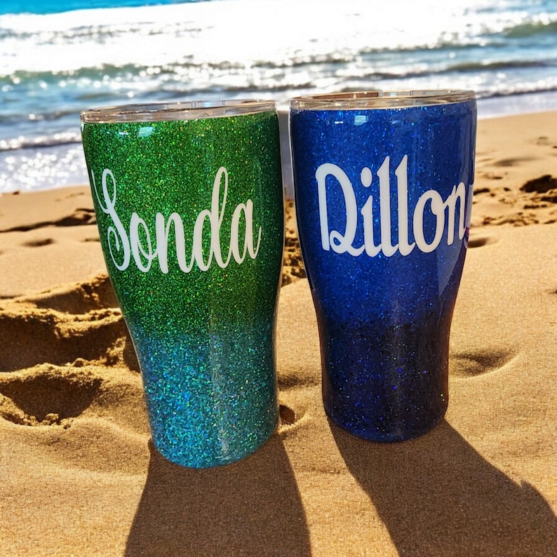 May include: Two personalized glitter tumblers with ombre colors. The tumbler on the left is green and blue with the name "Sonda" on it. The tumbler on the right is blue with the name "Dillon" on it.