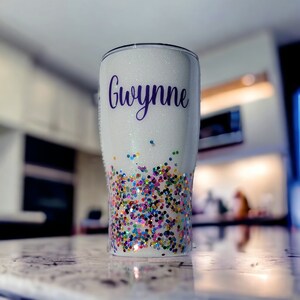 May include: A white glitter tumbler with the name "Gwynne" in purple script. The bottom half of the tumbler is decorated with colorful confetti. The tumbler is designed to keep drinks cold or hot.