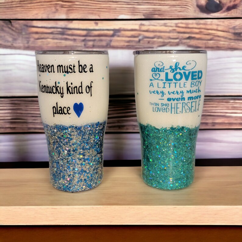 May include: Two white tumblers with blue glitter and text. The tumbler on the left says "Heaven must be a Kentucky kind of place" with a blue heart. The tumbler on the right says "and she LOVED A LITTLE BOY very, very much - even more than she LOVED HERSELF".