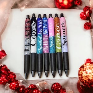May include: Seven glitter pens with black grips are displayed on a white surface. Each pen has a different color and text: "It Is What It Is Cows", "Spoiled Ass Wife", "It's Ok To Not Be Like", "Spoiled Wife", "Grace", and "Spencer". Festive decorations surround the pens.