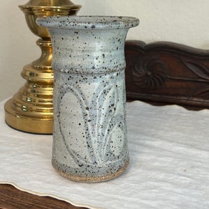 Puede incluir: Un jarrón de cerámica azul claro moteado con un borde acampanado y una superficie texturizada. El jarrón presenta un diseño en forma de hoja y mide aproximadamente 15 cm de altura. La base es de color natural sin esmaltar. Una lámpara de latón está en el fondo.