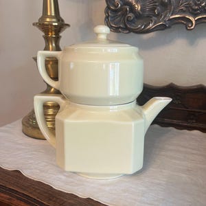 May include: A vintage, cream-colored teapot with a matching lid. The teapot has a geometric, octagonal base and a rounded top. It features a spout and a handle on each section. The teapot is sitting on a white, textured surface.